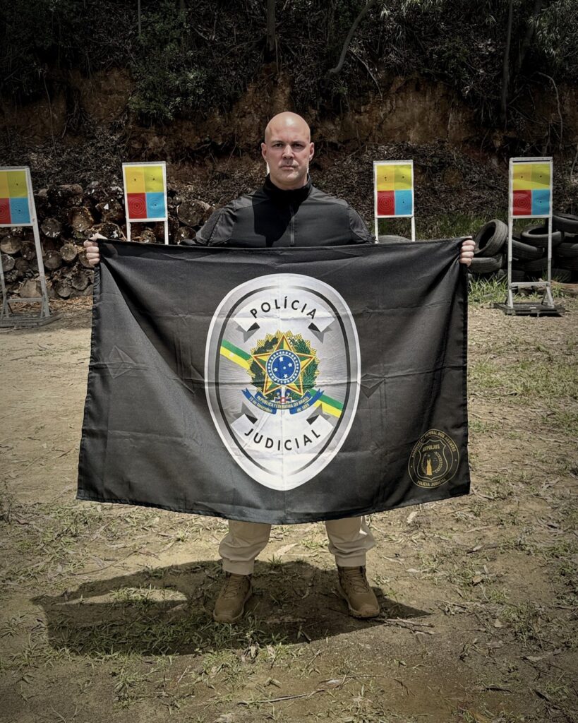 Prof. Alessandro Marques, Agente de Polícia Judicial, segurando a bandeira da Polícia Judicial em um estande de tiro